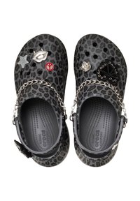 Schwarze Clogs mit Leopardenmuster und Kettenakzenten, verziert mit verschiedenen dekorativen Charms wie einem Stern, Lippen, einem Herzen und einer Rose, auf einer strukturierten Oberfläche.