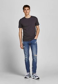 Sötétszürke rövid ujjú póló szakadt kék farmerrel párosítva. Fehér és fekete tornacipőben viselve, amely színes részletekkel rendelkezik.