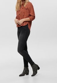 Rostfarbendes Hemd mit Knopfleiste, schwarze enge Jeans und schwarze Ankle-Boots mit Schlangenprägung und niedrigem Blockabsatz. Einfaches und modernes Design.