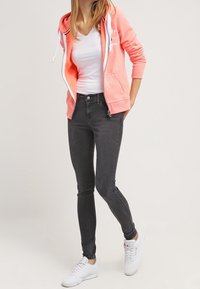 Donna che indossa una felpa corallo con zip, una maglietta bianca a V, jeans skinny neri e sneakers bianche, in piedi con una mano dietro la schiena.