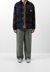Übergroße marineblaue Jacke mit mehrfarbigen Streifen, ausgestattet mit einer Knopfverschluss und Tasche, getragen über einem beigen Hemd und lockeren grünen Hosen.