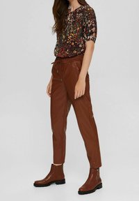 Bruine faux leren joggers met elastische taille, gecombineerd met een kortemouwen bloes in donkere tinten en bruine enkellaarsjes met een zijrits.