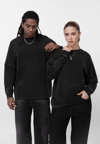 Sudaderas negras de gran tamaño con mangas raglán y detalles de costuras visibles, combinadas con pantalones de mezclilla oscuros, llevadas por dos modelos.