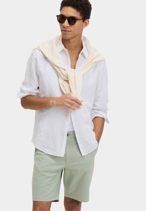 Camisa blanca abotonada, pantalones cortos verde claro y suéter color crema sobre los hombros; gafas de sol completan el look. Atuendo casual pero coordinado.