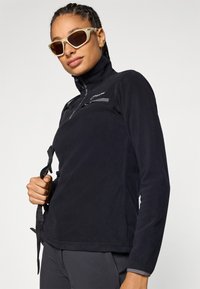 Pullover in pile nero con colletto alto, tasca sul petto con zip e dettagli grigi. Il modello indossa occhiali da sole chiari e tiene una tracolla.