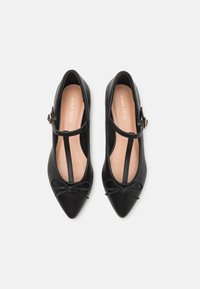 Bailarinas negras de punta afilada con diseño de tira en T, acento de lazo en la puntera y acabado de cuero suave. El interior cuenta con un forro beige.