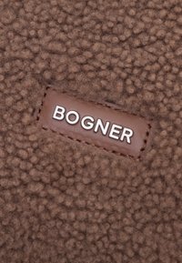 Brązowy polarowy materiał o teksturowanym wykończeniu z prostokątną skórzaną łatą z wytłoczonym w srebrnym letteringiem nazwiskiem marki "BOGNER".