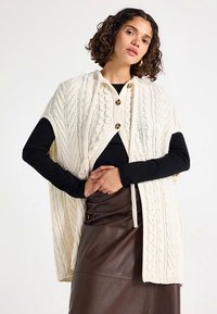 Kréčikový smotanový pončo s golierom, zdobený zlatými gombíkmi, s otvorenými stranami a textúrovaným vzorom. Nosený cez čierne tričko s dlhým rukávom.