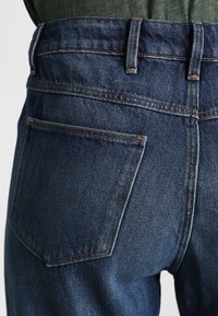 Vaqueros de mezclilla azul oscuro con cintura alta, que cuentan con un solo bolsillo trasero y detalles de costura visibles. Textura suave y corte clásico.