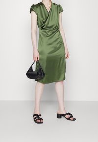 Robe en satin vert avec des manches courtes, un décolleté plissé, coupe au niveau du genou ; associée à un sac à main noir et des sandales à talons avec des lanières noires.
