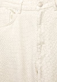 Detailný záber na béžové textúrované nohavice s pútkami na opasok, prednou vreckom a kovovým gombíkom s gravírovaním "PULL&BEAR."