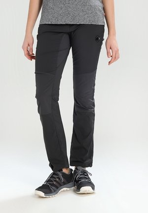 Person iført sorte patchwork vandrebukser, grå t-shirt og sorte trail løbesko står på hvid baggrund.