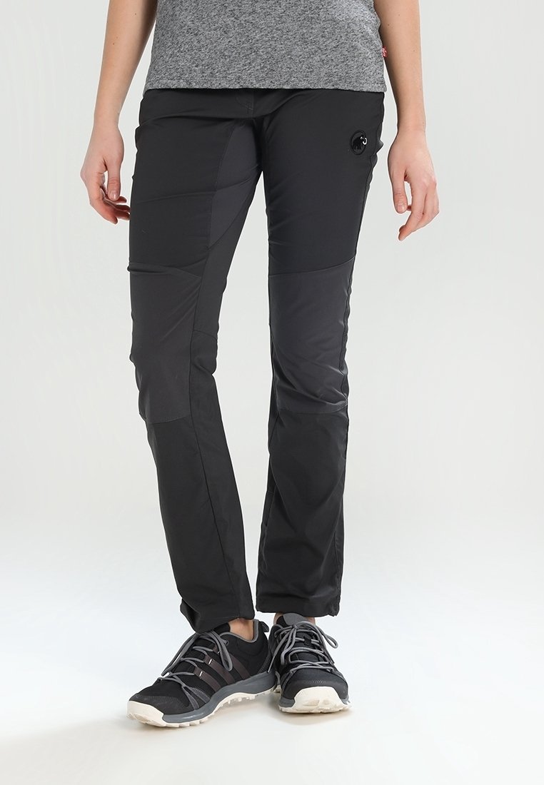Personne portant un pantalon de randonnée noir patchwork, un t-shirt gris et des chaussures de trail noires, debout sur fond blanc.