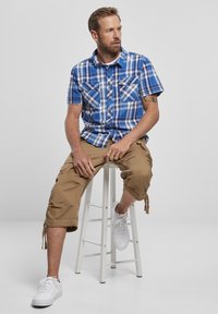 Kék kockás, rövid ujjú ing, két mellzsebbel, bézs cargo sorttal párosítva. A modell fehér sportcipőt visel, miközben egy széken ül.