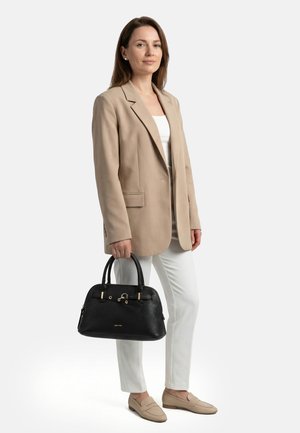 Woman wearing beige blazer, white top, and white pants holding a black handbag, standing against a plain white background.