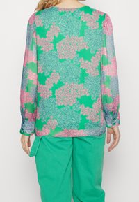 Blusa floreale verde con maniche lunghe, caratterizzata da un motivo di fiori rosa e blu, realizzata in tessuto leggero e trasparente.