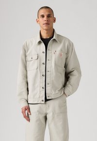 Chaqueta de denim beige claro con un corte relajado, que cuenta con dos bolsillos en el pecho, cierre de botón y un sutil parche de logo en el bolsillo izquierdo.