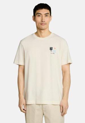 Mężczyzna noszący jasnobeżowy t-shirt z krótkim rękawem z małą grafiką parasoli i tekstem na lewej piersi, w połączeniu z beżowymi spodniami.