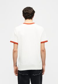 Άτομο με κοντά σκούρα μαλλιά που φορεί λευκό T-shirt με πορτοκαλί τρέσα στον γιακά και στα μανίκια, στέκεται πλάτη σε απλό φόντο.