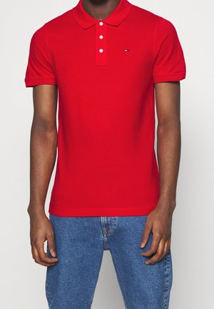 Man draagt een felrode korte mouw polo met knopen en blauwe spijkerbroek, staand tegen een effen lichte achtergrond.