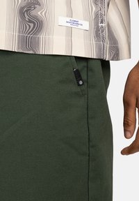 Short-sleeve shirt with beige and gray wavy pattern, paired with green pants featuring a side pocket and a black brand tag.