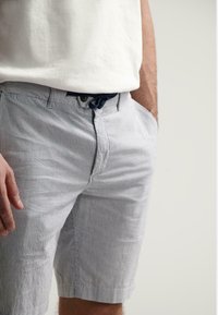 Shorts rayés en bleu clair et blanc, fabriqués en tissu doux. Présente une fermeture boutonnée, des passants de ceinture et des poches latérales.