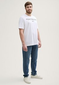 Camiseta de algodón blanca de manga corta con un diseño de texto en negro, combinada con unos vaqueros de mezclilla azules y zapatillas blancas, de pie contra un fondo neutro.
