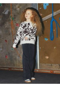 Jeans neri con borchie argentate lungo l'orlo, abbinati a un maglione a stampa mucca nei toni del beige e del marrone. Capelli lunghi e ricci di colore biondo. Sfondo di una parete da arrampicata.