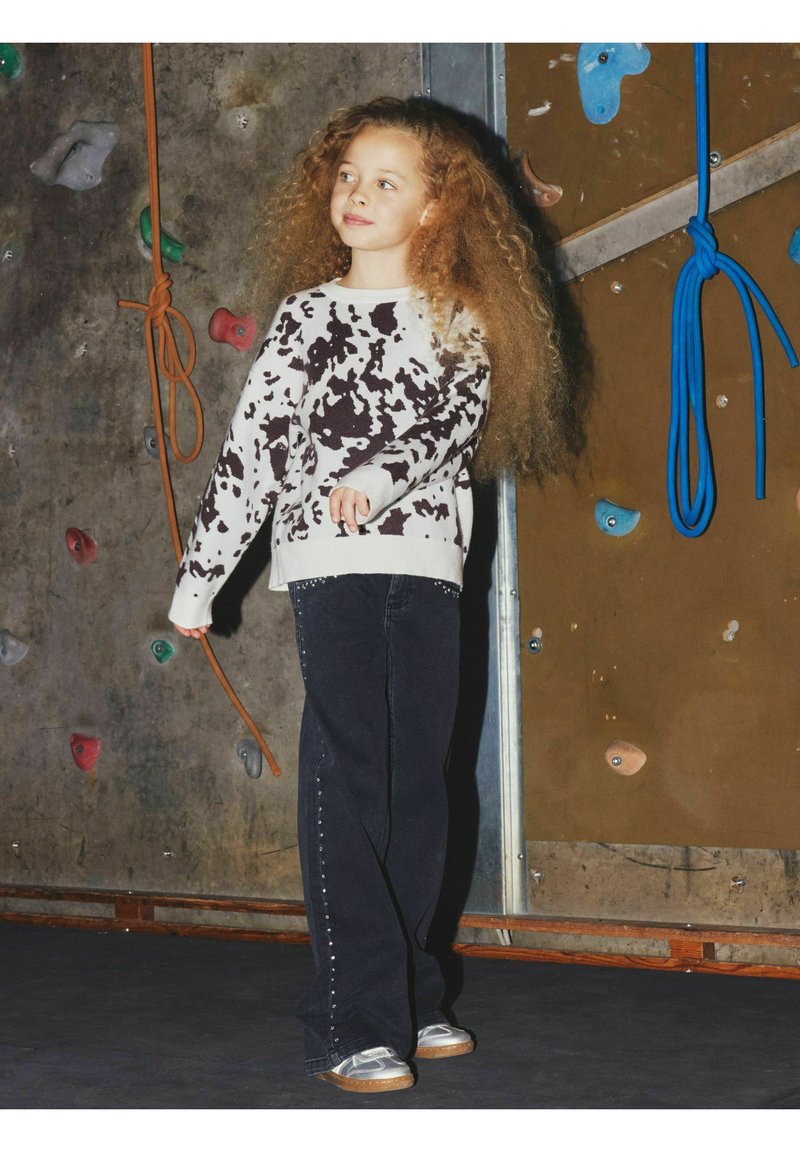 Jeans neri con borchie argentate lungo l'orlo, abbinati a un maglione a stampa mucca nei toni del beige e del marrone. Capelli lunghi e ricci di colore biondo. Sfondo di una parete da arrampicata.