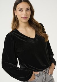 Schwarzes Samtoberteil mit V-Ausschnitt und puffierten langen Ärmeln. Verfügt über eine glatte Textur und ein tailliertes Design. Kombiniert mit hochgeschnittenen grauen Jeans.