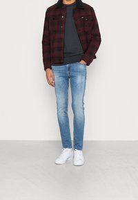 Chaqueta a cuadros roja y negra con un collar negro, camisa gris debajo, combinada con jeans ajustados de color azul claro y zapatillas blancas.