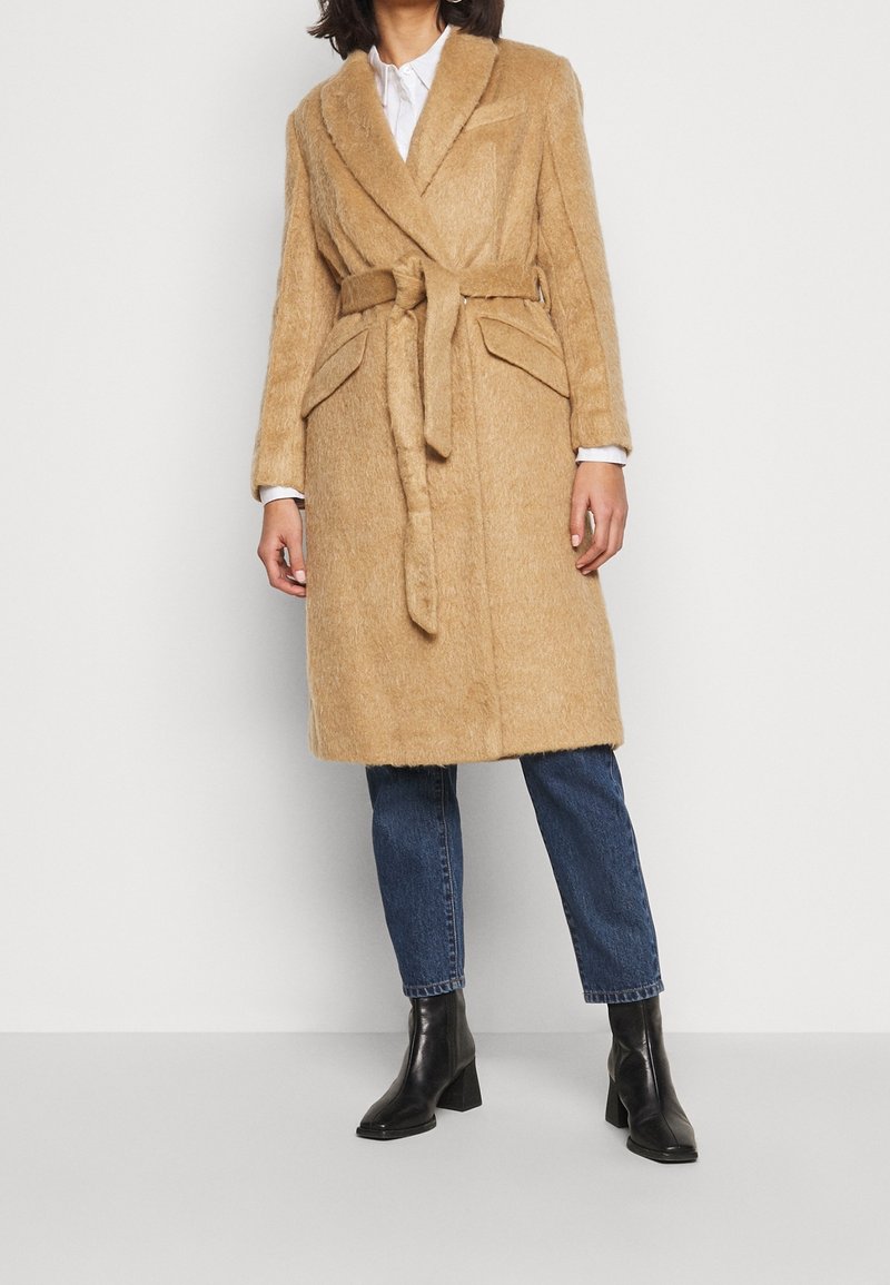 Femme portant un manteau en laine camel ceinturé longueur genoux, par-dessus une chemise blanche, un jean bleu et des bottines noires, sur un fond uni.