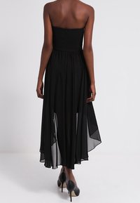 Černé bezramínkové šaty s volánkovou, průhlednou sukní a asymetrickým lemem. Hladká tkanina a nařasený pas pro jemný splývavý efekt.