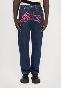 Jeans en denim bleu foncé avec une coupe décontractée, présentant un grand graphique de chien cartoon rose sur la cuisse gauche. Des baskets noires complètent le look.