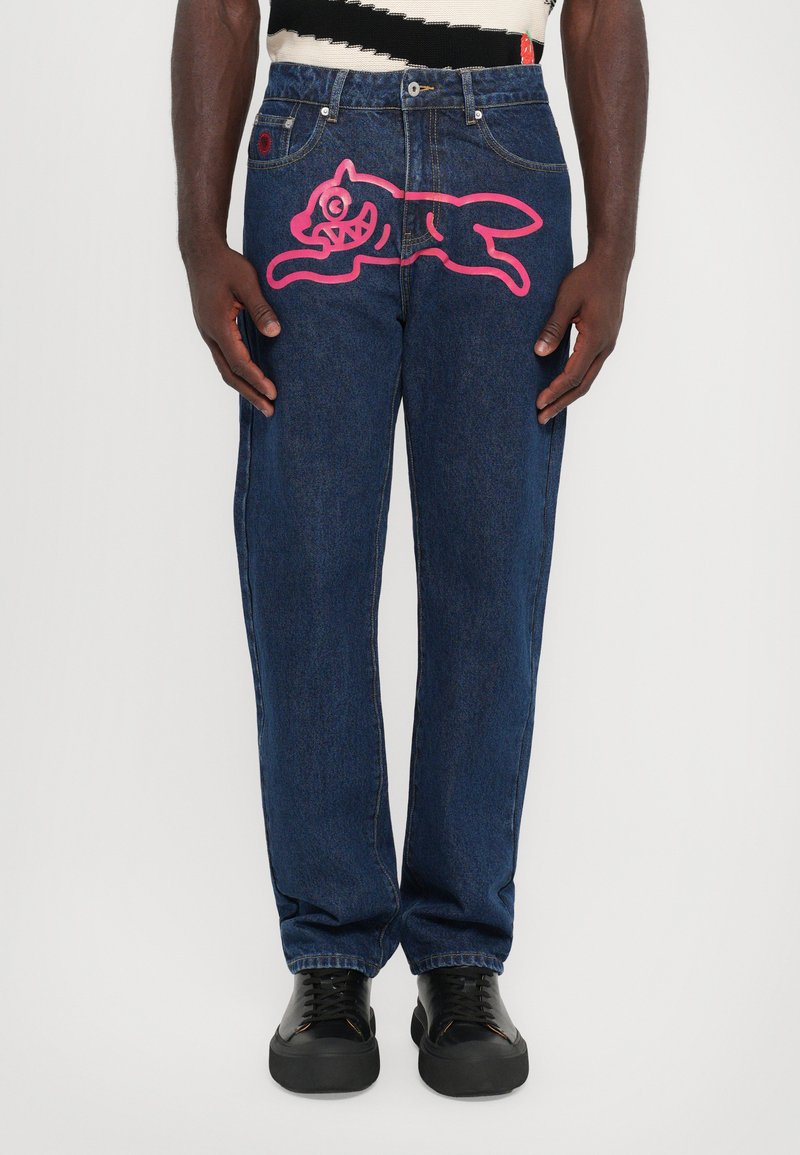 Jeans en denim bleu foncé avec une coupe décontractée, présentant un grand graphique de chien cartoon rose sur la cuisse gauche. Des baskets noires complètent le look.