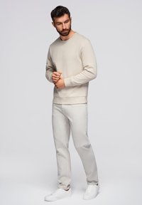 Felpa beige chiaro con colletto a girocollo, polsini e orlo a coste; abbinata a pantaloni grigi chiari e sneakers bianche. Tessuto con texture liscia.