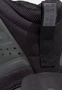Close-up van de hiel van een zwarte sneaker met mesh-stof, een trekkoordje met het label "Swiss Design" en een geperforeerd detail van een synthetische zool.