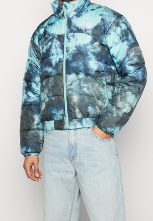 Man in een blauw en zwart tie-dye pufferjack met het logo van "The North Face", lichte spijkerbroek, met één hand in de zak, staand tegen een witte achtergrond.
