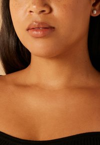 Nahaufnahme des Halses und des unteren Gesichtes einer Person mit glatter Haut, hellbraunen Tönen und kleinen Ohrsteckern mit rechteckigem Design.