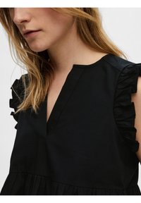 Svart textil topp med v-ringning och volangaccent vid axlarna. Den nedre delen är samlad, vilket skapar en strömlinjeformad effekt.