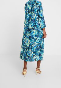 Robe florale avec des manches trois-quarts, fond bleu profond, et roses jaunes et turquoise éclatantes. Mi-longue avec un tissu fluide.