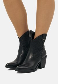 Botines de cuero negro hasta el tobillo con punta afilada, costuras decorativas, cremallera lateral y tacón ancho. Detalles texturizados en los laterales.