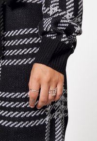 Indumento lavorato a maglia a righe bianche e nere con polsini a coste, indossato su una mano che mostra anelli d'argento con design a onde e un tatuaggio.