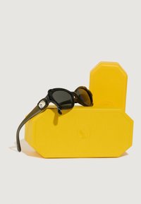 Gafas de sol con lentes oscuros y montura negra, descansando sobre un estuche octagonal texturizado de color amarillo. Detalle de hardware notable en la montura.