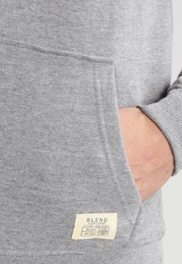 Grå hettegenser med frontlomme, teksturert stoff og etikett med "BLEND"-tekst og produktdetaljer.