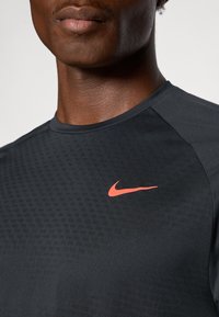 Czarna koszulka sportowa Nike z teksturowanym wzorem i małym, jaskrawopomarańczowym logo swoosh na klatce piersiowej. Krótkie rękawy, okrągły dekolt.