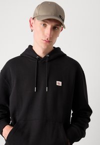 Zwarte katoenen hoodie met een voorzak en zilveren trekkoorden, voorzien van een klein geborduurd logo. In combinatie met een lichtgrijze pet.