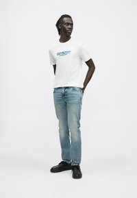 Camiseta blanca de algodón con texto gráfico azul, combinada con jeans de mezclilla azul claro y zapatos negros. El modelo está de pie de manera casual con las manos en los bolsillos.