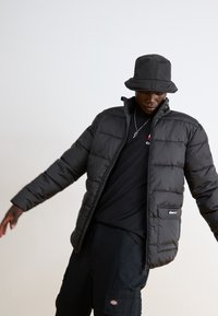 Giubbotto in piuma nero con design trapuntato, colletto alto e chiusura con zip. Indossato sopra una maglietta nera, abbinato a un cappello a secchiello nero.