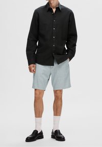 Camicia nera con bottoni e maniche lunghe, abbinata a pantaloni corti azzurri. Mocassini neri e calzini bianchi completano l'outfit.